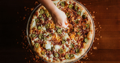 A hand sprinkles toppings on a pizza loaded with cheese, jalapeños, and crispy bits, resting on a wooden table.