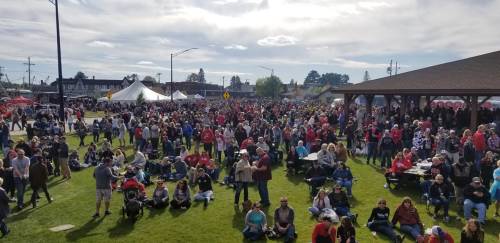 A large crowd gathers at an outdoor event, with tents and a pavilion under a partly cloudy sky.