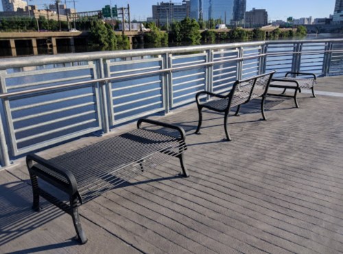 Three metal benches on a wooden deck overlooking a calm river, with city buildings in the background.