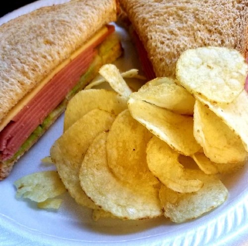 A sandwich with layers of ham and cheese on whole grain bread, served with a side of potato chips.
