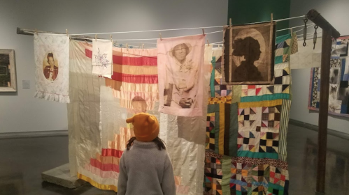 A child in a yellow hat stands before a colorful quilt display featuring various fabric patterns and portraits.