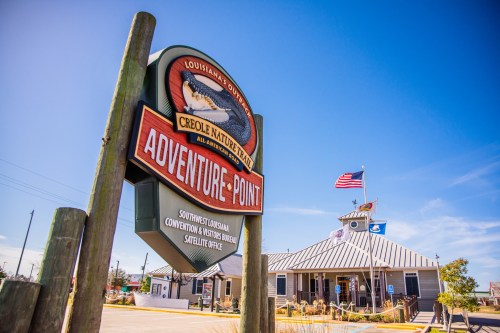 Sign for Adventure Point at Creole Nature Trail, with a building and American flag in the background under a clear blue sky.