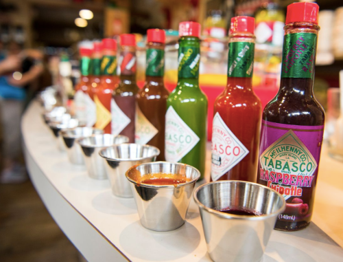 A row of colorful Tabasco sauce bottles with small metal cups for tasting in a vibrant store setting.
