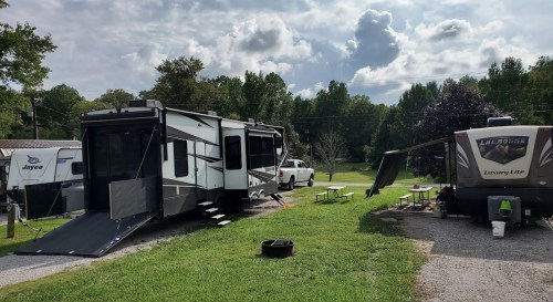 Two RVs parked in a grassy area with picnic tables, surrounded by trees and a cloudy sky.