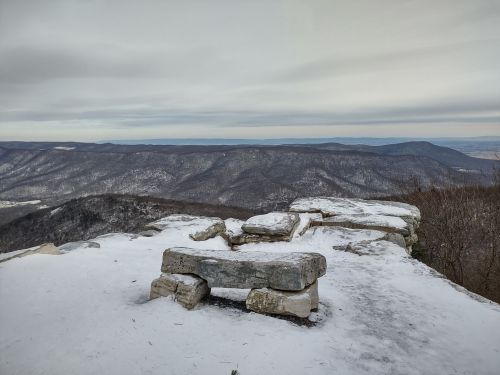 A snowy mountain overlook with a stone bench, showcasing distant hills under a cloudy sky.