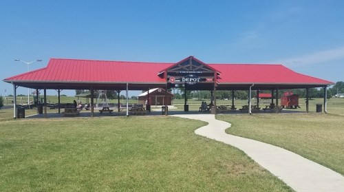 A large pavilion with a red roof in a grassy park, featuring picnic tables and a pathway leading to it.