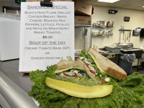 A sandwich with grilled chicken, Swiss cheese, and veggies on wheat bread, next to a pickle and a sign for daily specials.