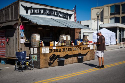 A rustic root beer stand with barrels and signage, featuring a man checking the menu in a sunny outdoor setting.