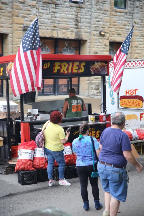 A food stall selling fries and sausages, with customers and American flags in a lively outdoor setting.