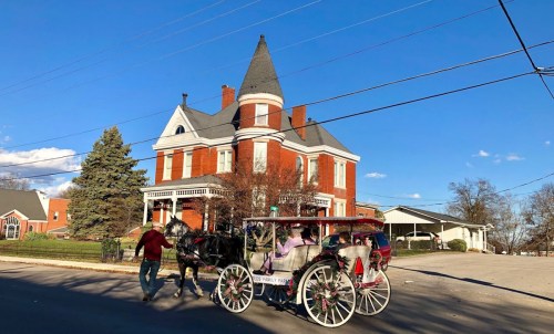 A horse-drawn carriage passes by a large, red brick Victorian house under a clear blue sky.