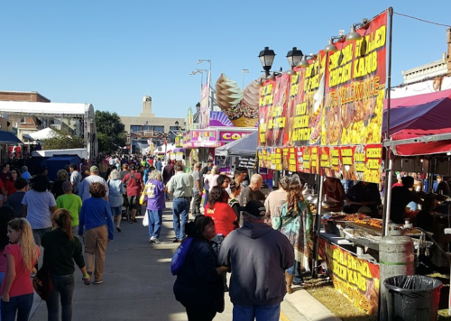 A bustling street fair with colorful food stalls and a crowd of people enjoying the festivities under a clear blue sky.