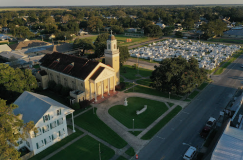 Aerial view of a church with a clock tower, surrounded by green lawns and a cemetery in the background.