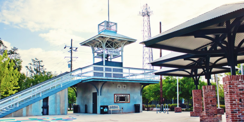 A modern train station with a tall platform, surrounded by greenery and a clear blue sky.