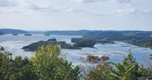 A scenic view of a river with islands, surrounded by lush greenery and rolling hills under a partly cloudy sky.