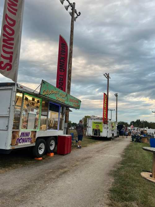 Food trucks lined up at a fair, with colorful banners and a cloudy sky in the background.