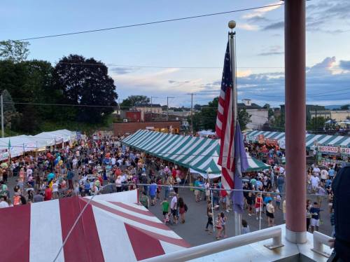 A bustling outdoor festival with crowds, colorful tents, and an American flag in the foreground under a cloudy sky.