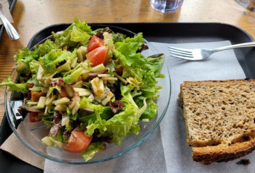 A fresh salad with greens, tomatoes, and apples, served alongside a slice of whole grain bread on a tray.