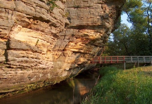 A rocky cliffside with layered stone formations beside a calm stream and a wooden walkway in a lush green setting.
