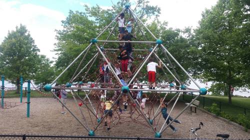 Children play on a large, geometric climbing structure in a park, surrounded by trees and a clear sky.