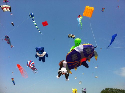 Colorful kites in various shapes, including animals and aliens, flying against a clear blue sky.