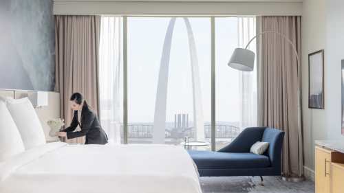 A hotel room with a bed and a view of a large arch structure through a window, featuring a woman arranging flowers.
