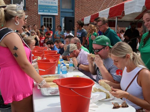 A crowd watches as contestants eat corn on the cob at a festival, with buckets and water bottles on the table.