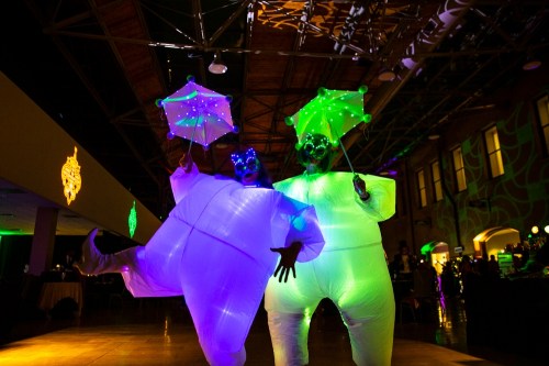 Two performers in glowing, inflatable costumes hold illuminated umbrellas, dancing in a vibrant, colorful venue.