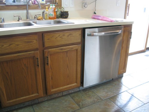 A kitchen with wooden cabinets and a stainless steel dishwasher next to a double sink on a tiled floor.