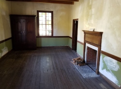 Empty room with wooden floors, a fireplace, and a dark wardrobe against the wall. Light streams through a window.