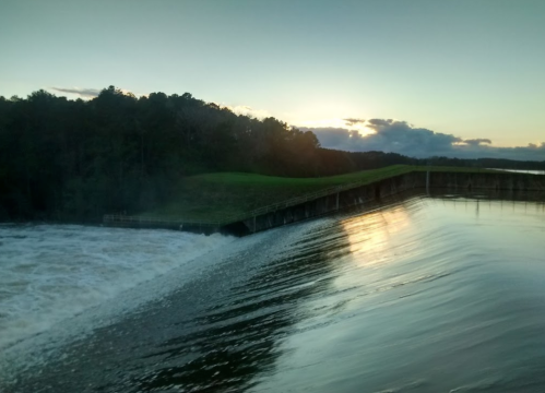A serene landscape featuring a dam releasing water at sunset, with trees and a calm sky in the background.