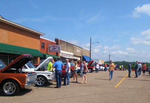 A street scene with classic cars on display and people mingling outside shops on a sunny day.
