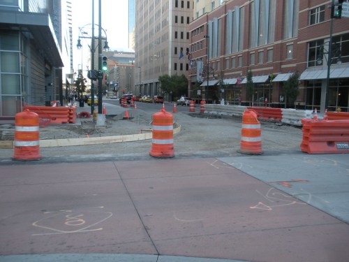 Construction site with orange traffic cones and barriers on a city street, surrounded by buildings and parked cars.