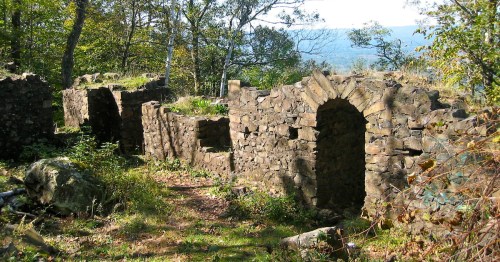Ruins of stone walls and arches surrounded by trees and greenery, with a scenic view in the background.