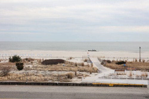 A serene beach scene with sandy dunes, a wooden walkway, and calm waters under a cloudy sky.