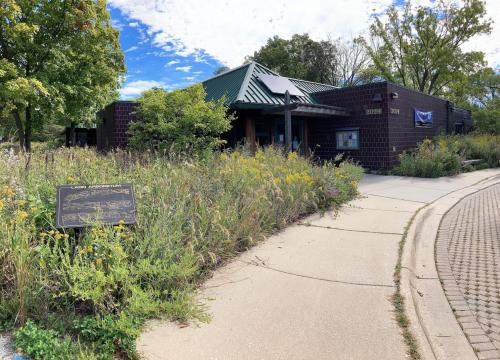 A nature center building surrounded by tall grasses and wildflowers, with a sign in front and a blue sky above.