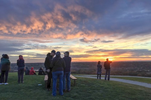 People gather on a hilltop, watching a vibrant sunset with colorful clouds in the sky.