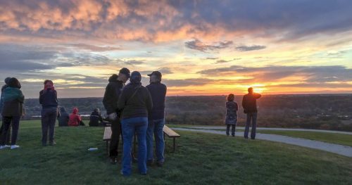 People gather on a hilltop, watching a vibrant sunset with colorful clouds in the sky.