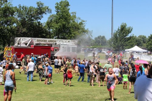 A crowd gathers at a park for a summer event, with a fire truck spraying water and people enjoying the festivities.