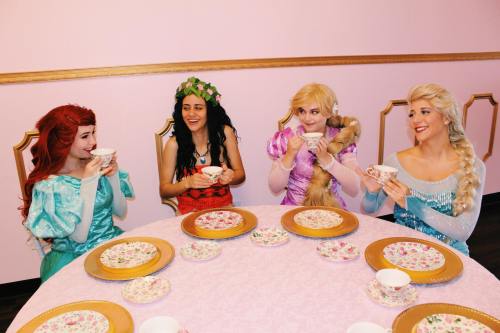 Four women dressed as princesses enjoy tea at a pink-themed table, smiling and holding teacups.