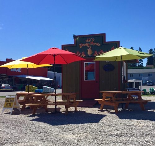 A colorful food truck with red, yellow, and green umbrellas, surrounded by picnic tables on a sunny day.