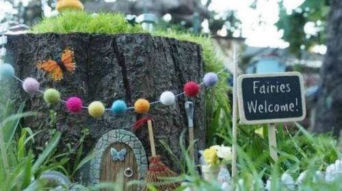 A whimsical fairy house on a tree stump, adorned with colorful decorations and a sign reading "Fairies Welcome!"