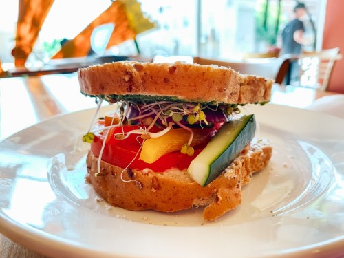 A colorful sandwich with layers of vegetables, sprouts, and whole grain bread, served on a white plate.
