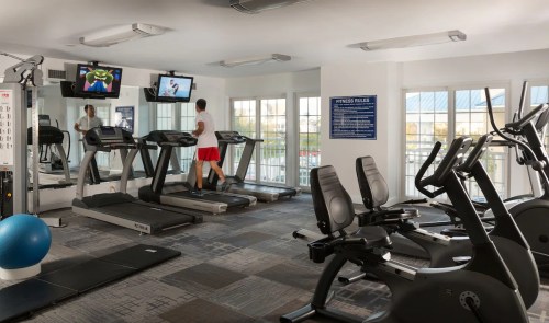 A modern gym with treadmills, exercise bikes, and a person jogging on a treadmill in front of two TVs.