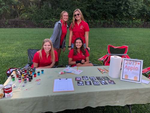 Four young women in red shirts pose at a table with craft supplies and a sign for an apple-themed event.