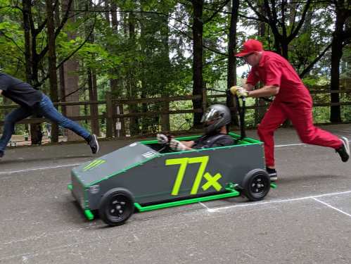 A person in a go-kart races down a track while another runs alongside, both surrounded by trees.