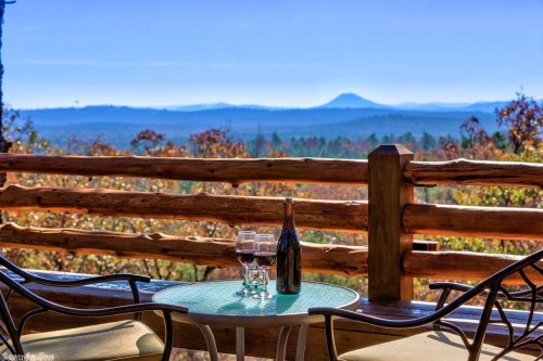 A scenic view from a balcony featuring two glasses of wine and a bottle, with mountains and autumn foliage in the background.