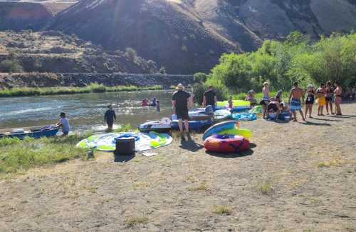 People enjoying a sunny day by the river, with inflatable rafts and boats along the shore. Greenery and hills in the background.