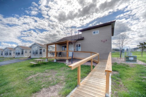 A modern house with a wooden ramp, surrounded by grassy areas and other similar homes under a cloudy sky.