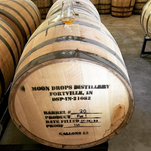 A wooden barrel labeled with "Moon Drops Distillery," containing a glass of amber liquid on top.