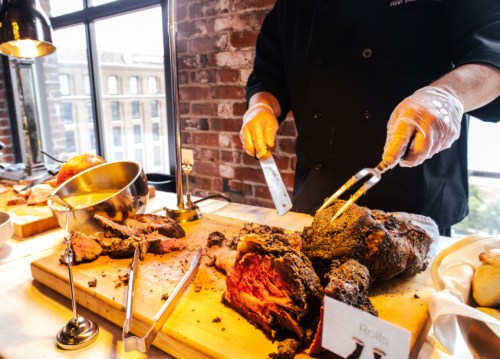 A chef carves a large roast on a wooden cutting board in a stylish setting with brick walls and natural light.